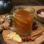 Segelas minuman bandrek khas Sunda hangat dengan jahe segar, kayu manis, bunga lawang, dan gula aren di atas meja kayu.
