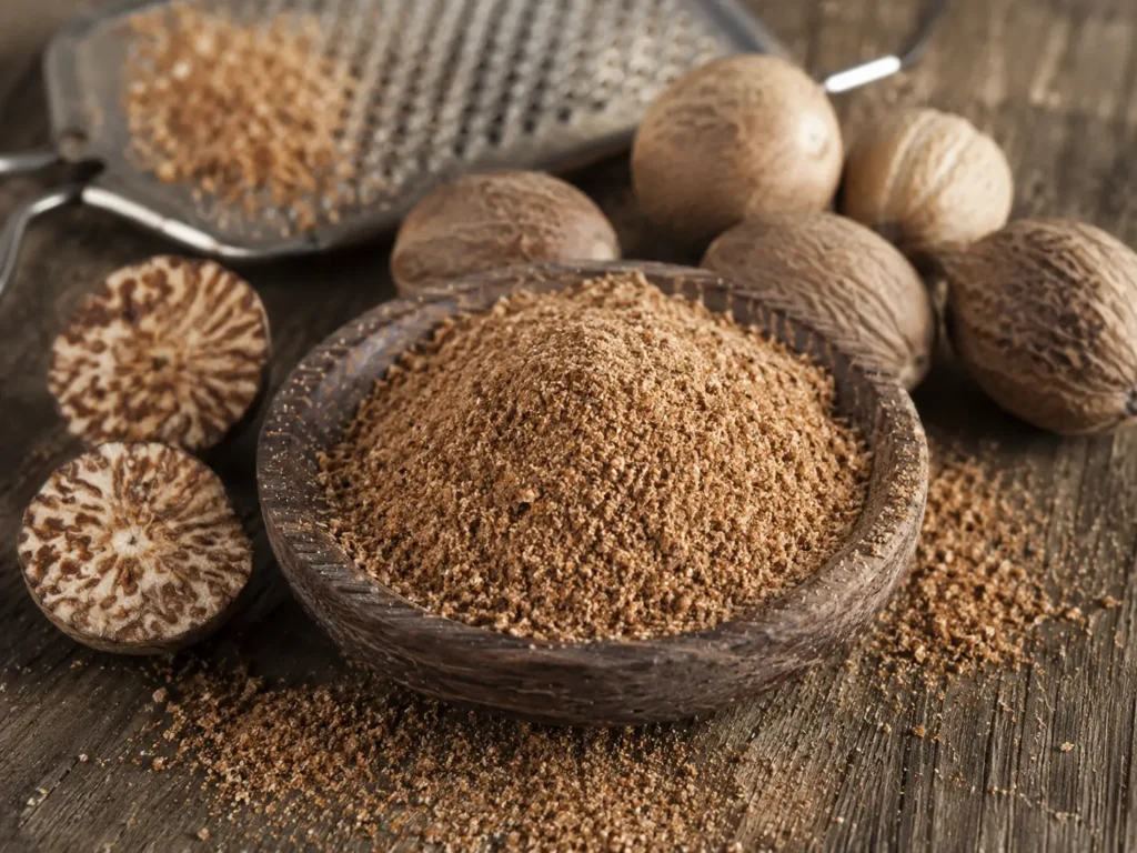 Bubuk pala halus dalam mangkuk kayu dengan biji pala utuh dan potongan biji pala sebagai rempah asli Indonesia.