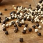Butiran lada hitam dan lada putih utuh bertebaran di atas meja kayu sebagai rempah dapur alami.