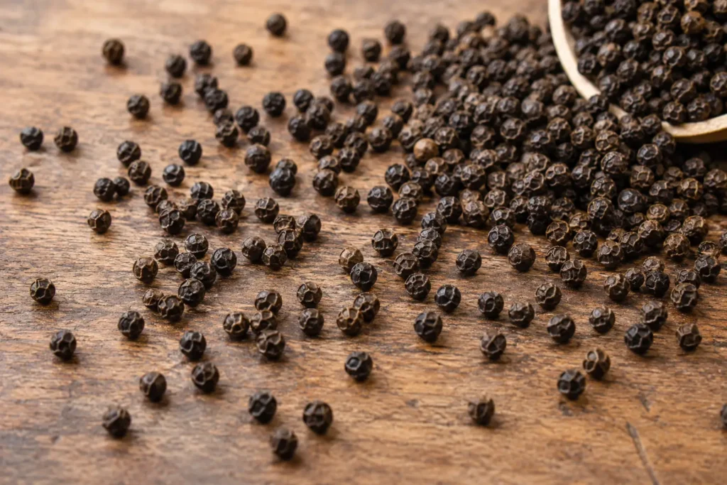 Butiran lada hitam utuh bertebaran di atas meja kayu dengan mangkuk berisi lada hitam sebagai rempah dapur.