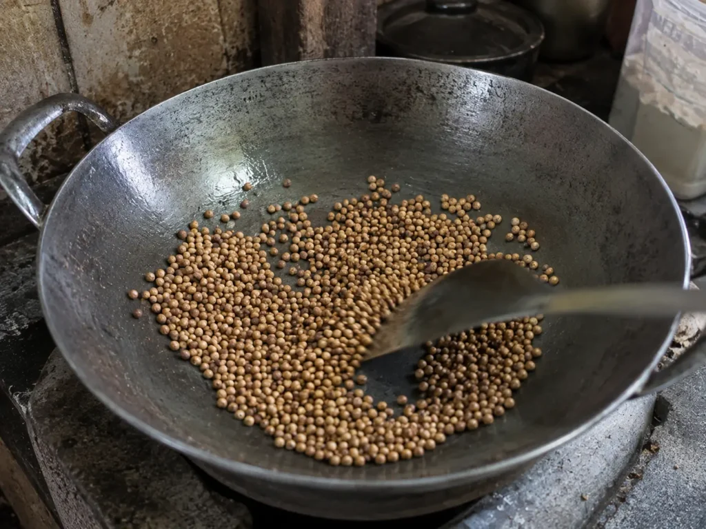 Biji ketumbar utuh sedang disangrai di dalam wajan besi menggunakan api kecil untuk mengeluarkan aroma rempah sebelum dihaluskan menjadi bumbu masakan.