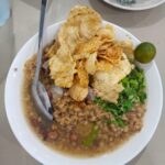 Bubur Pekong khas Singkawang dengan daging sapi, kerupuk goreng, kacang, daun bawang, dan jeruk limau dalam kuah kaldu gurih